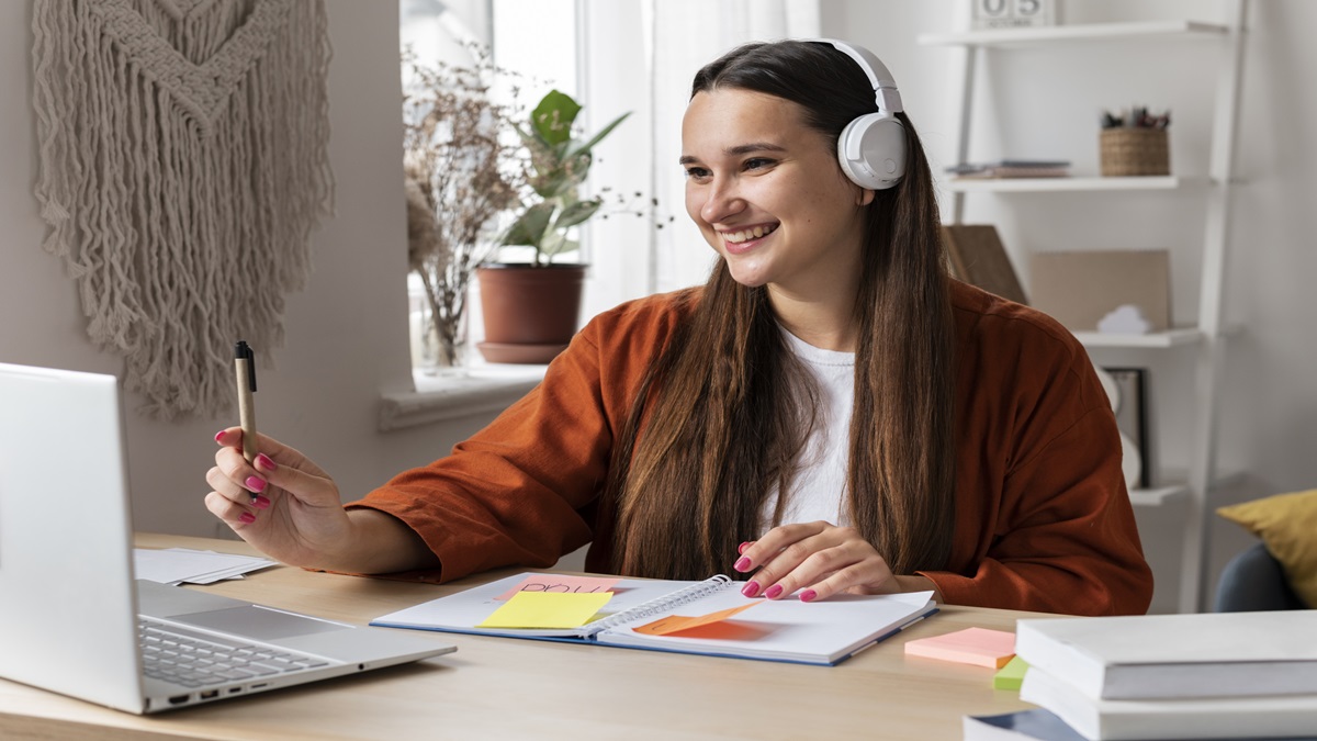 Pessoa estudando com fones de ouvido, laptop e caderno sobre a mesa